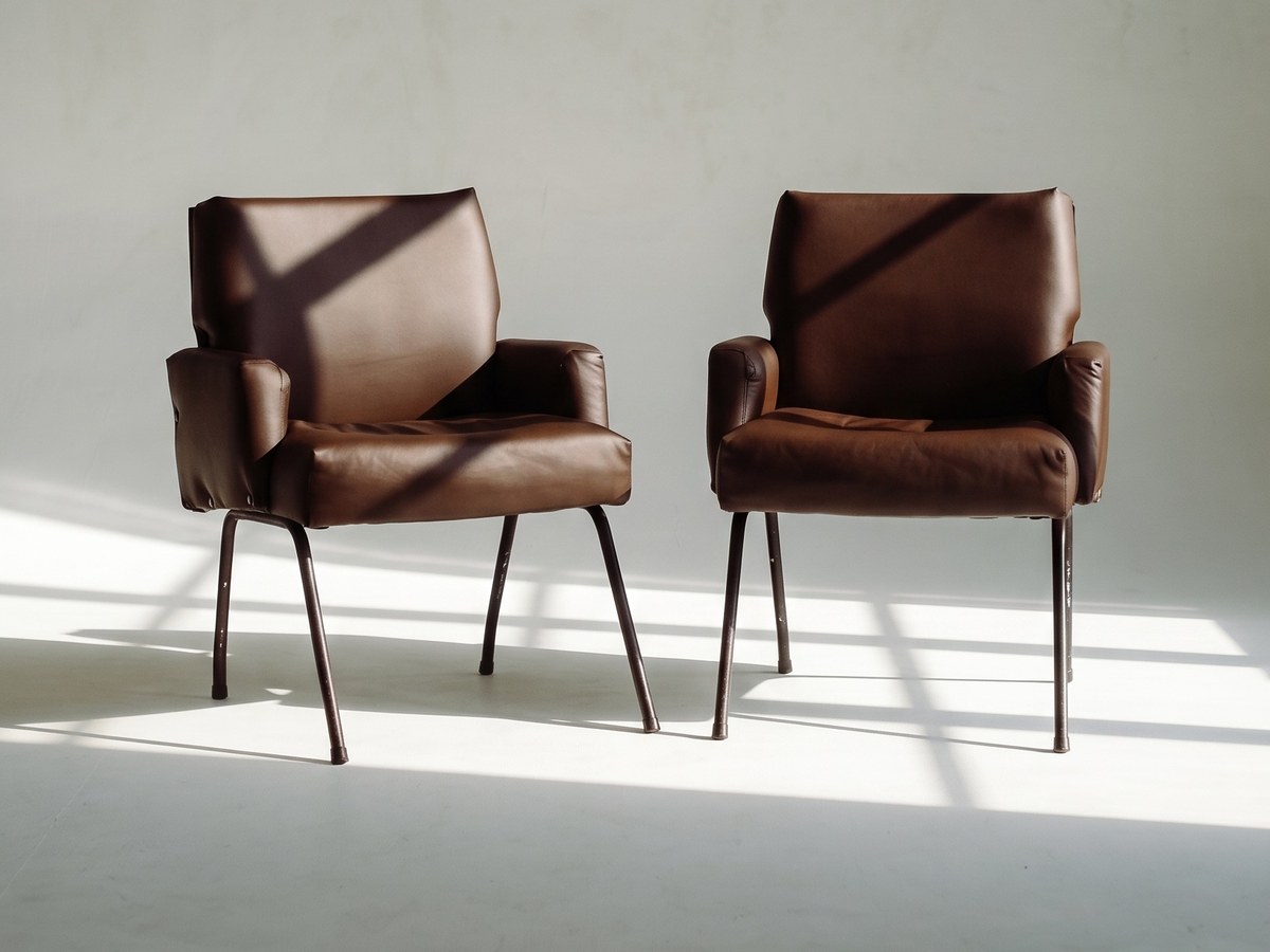 Two brown leather armchairs side by side on a concrete floor, lit by a window